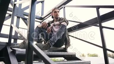 一个英俊的家伙，手里拿着智能<strong>手机</strong>，坐在<strong>日</strong>落时分一座工业<strong>建筑</strong>的楼梯上。 年轻的嬉皮士